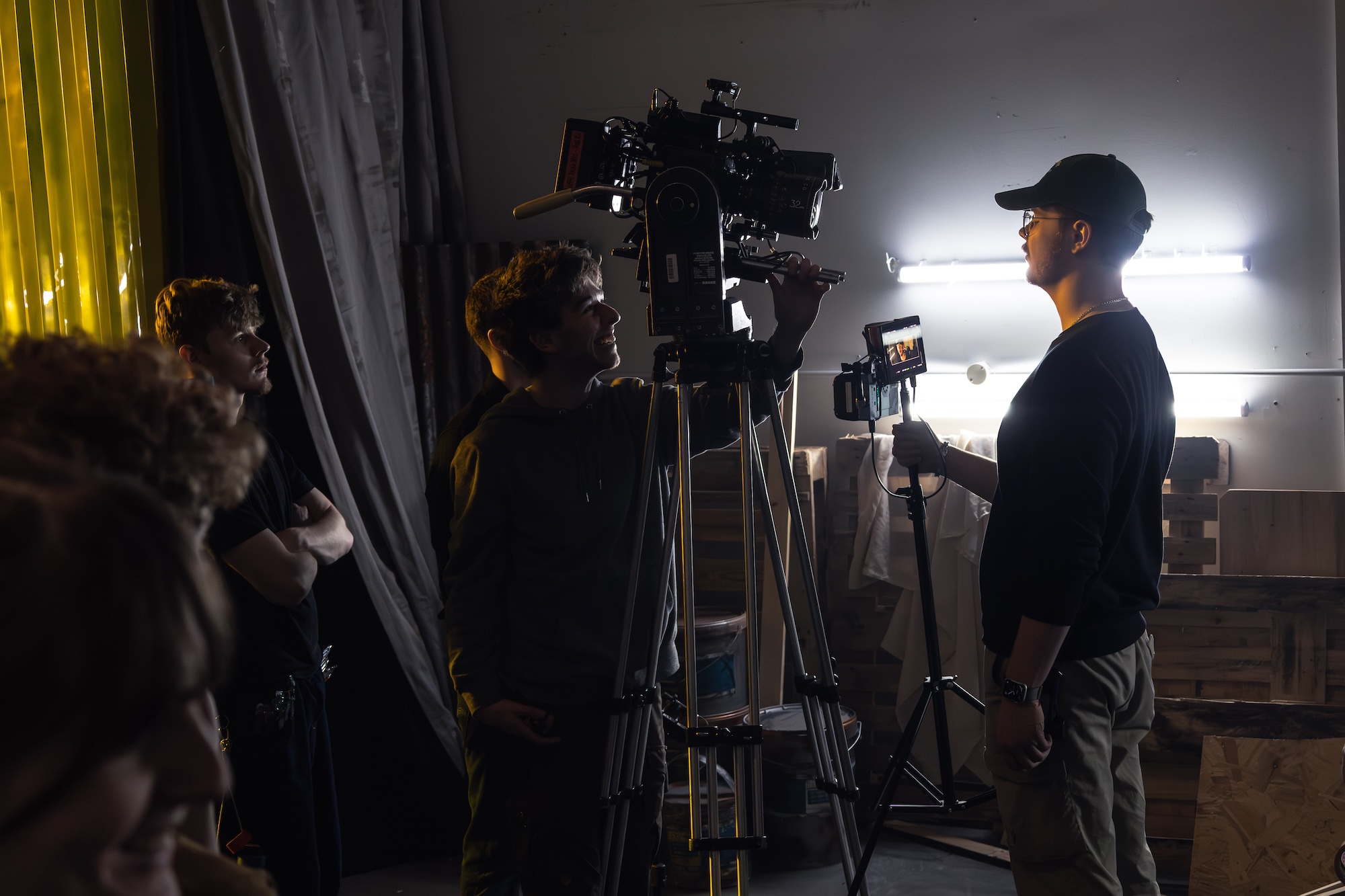 A film crew is working in a dimly lit studio. One man adjusts a camera mounted on a tripod, while another man holds a monitor displaying the camera feed. Two crew members observe in the background, surrounded by various studio equipment.