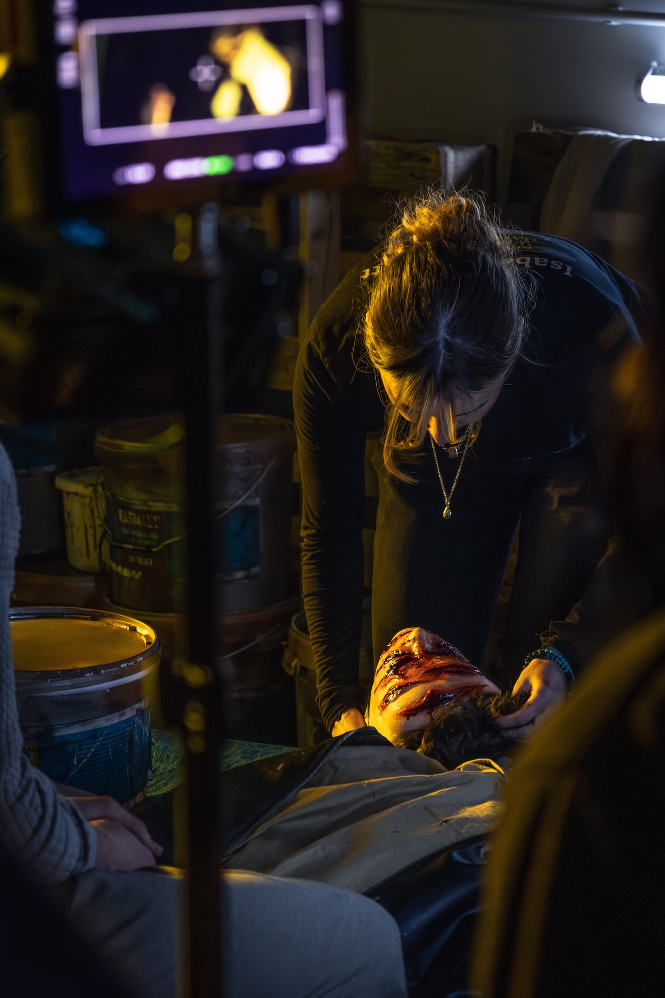 A woman wearing a black outfit leans over a person lying on the ground with visible wounds on their head. The setting includes containers in the background and soft lighting, indicating a staged scene related to an emergency.