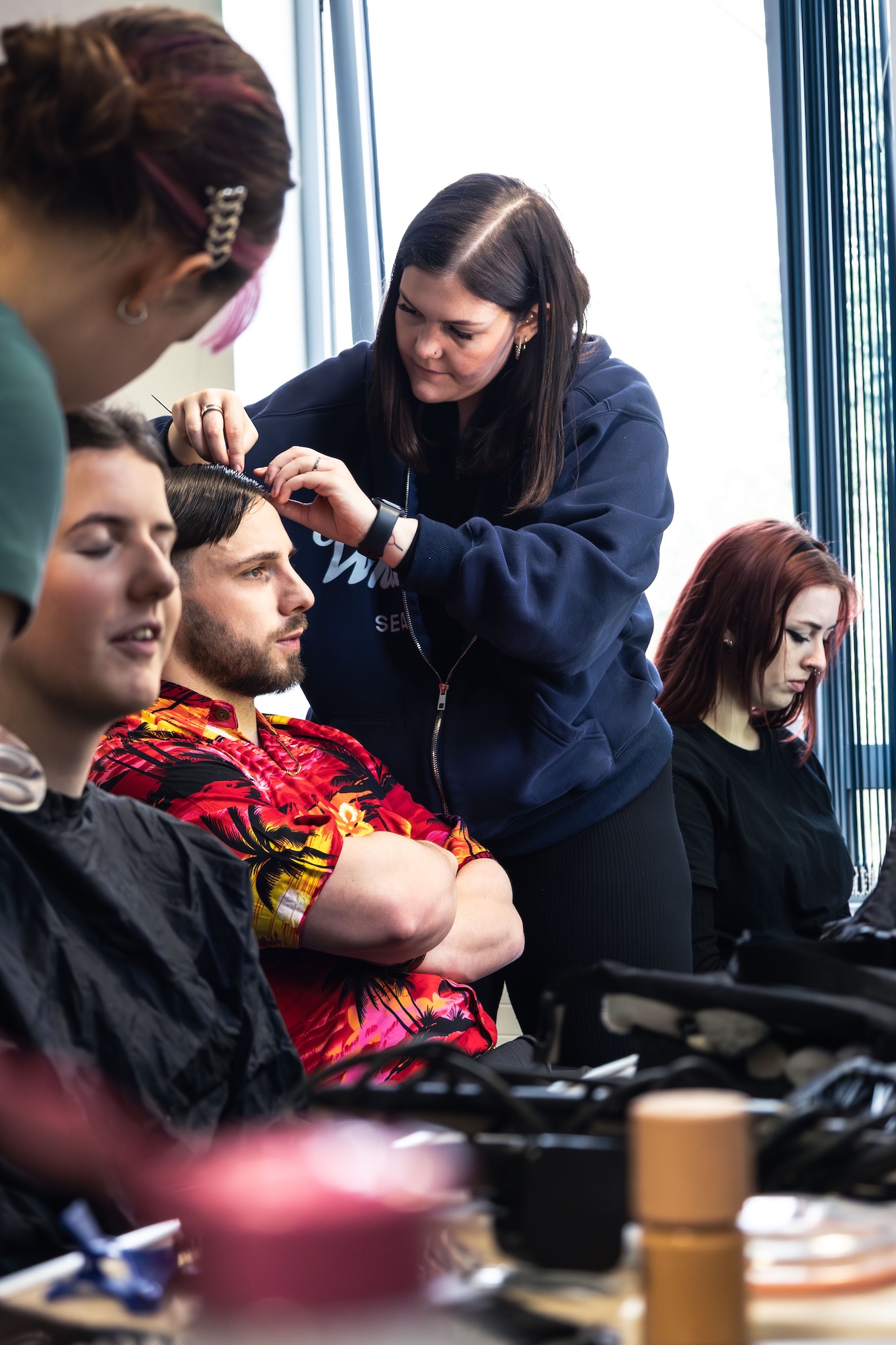 A hairdresser, dressed in a navy hoodie, styles the hair of a young man wearing a floral shirt, who is sitting with his arms crossed. Nearby, two women are seated, one with red hair and the other with dark hair, both in salon capes.