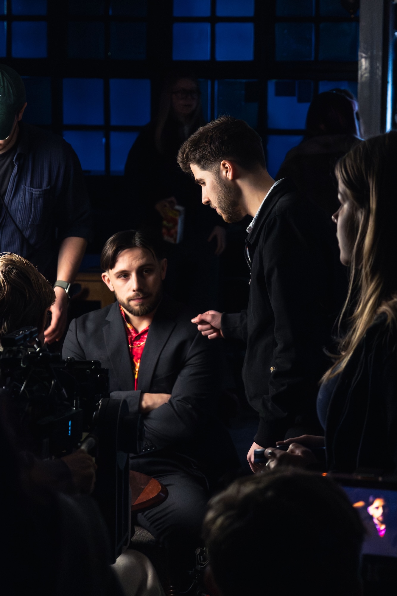 A man in a black suit and red shirt sits at a table, while another man in a black jacket leans in to speak to him. The background shows a dimly lit room with crew members visible, indicating a filming or production setting.