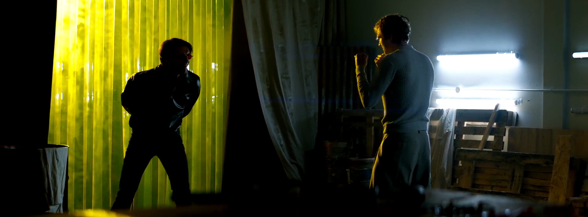 Two men are positioned facing each other in a dimly lit space. One man is wearing a black jacket and appears to be in a defensive stance, while the other, dressed in a grey outfit, is in a fighting position. Yellow transparent curtains are visible in the background.