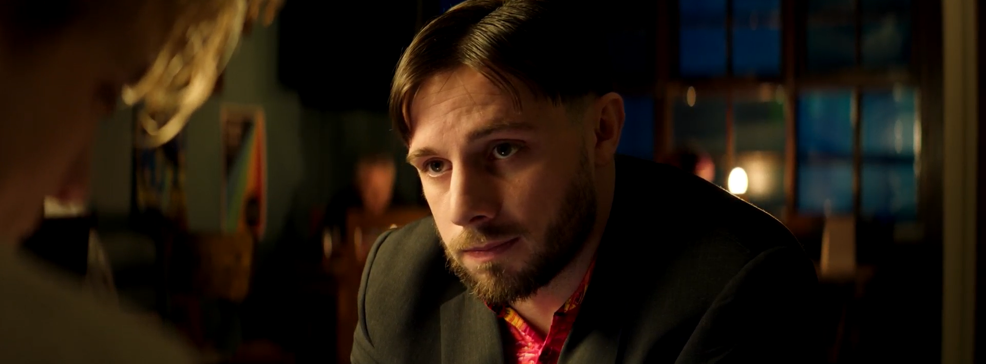 A man with short brown hair and a beard wears a dark suit and pink shirt while sitting at a table. The background is dimly lit with a few people visible and large windows showing a night scene.