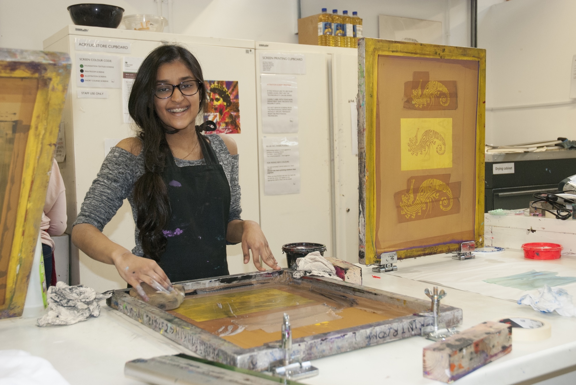 Young person wearing green apron works at printmaking table with screen printing frame in art studio.