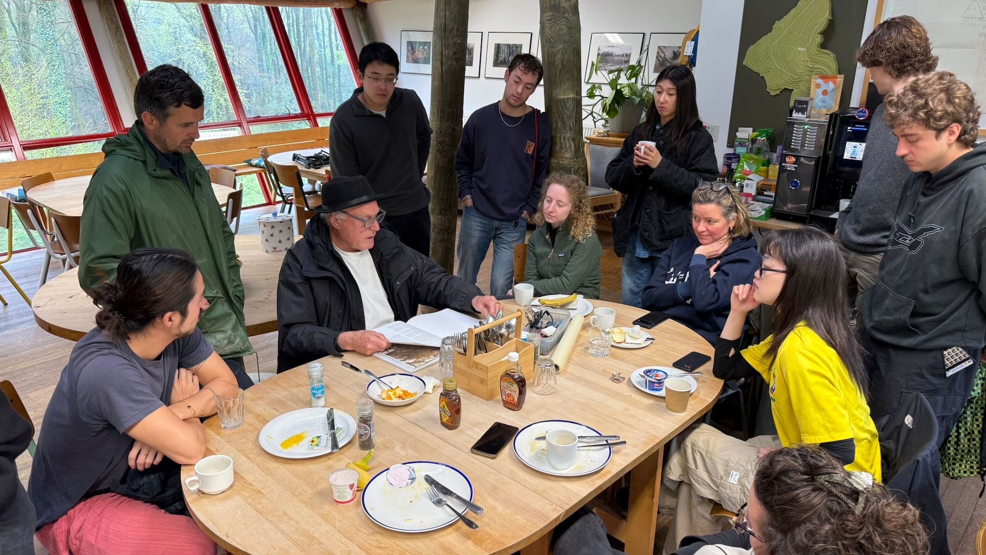 A group of people gathered around a wooden table during a discussion, with cups, plates and papers spread across it in a bright room with large windows.