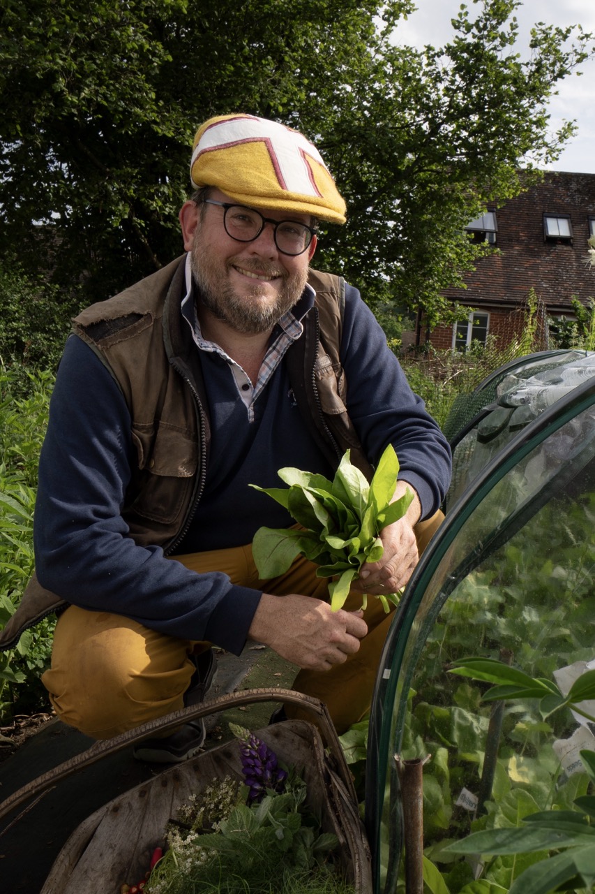 A smiling man wearing a yellow cap and glasses kneels beside a green glass greenhouse, holding a small potted plant in a garden.