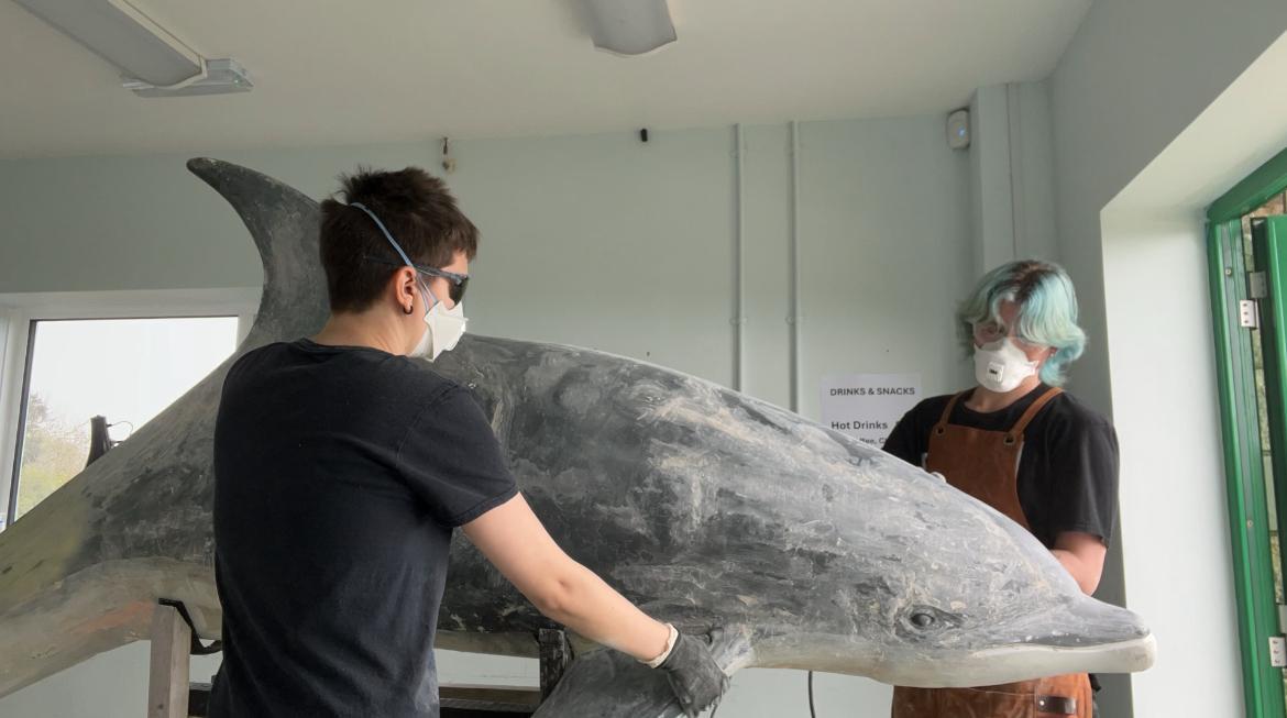 Two BA (Hons) Modelmaking students in masks examining a large grey dolphin statue in a white studio room at Durlston Country Park.