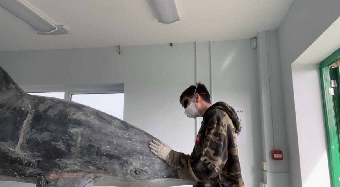 A BA (Hons) Modelmaking student in a camouflage jacket and white mask working on a large dolphin sculpture in a white-walled room with ceiling lights.