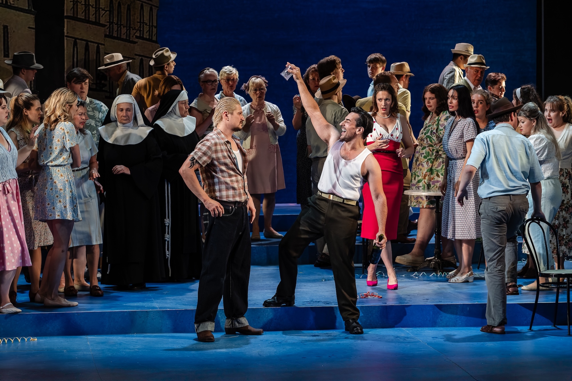 Opera performance with ensemble cast on blue-lit stage, featuring performers in period costumes including nuns' habits and formal wear.