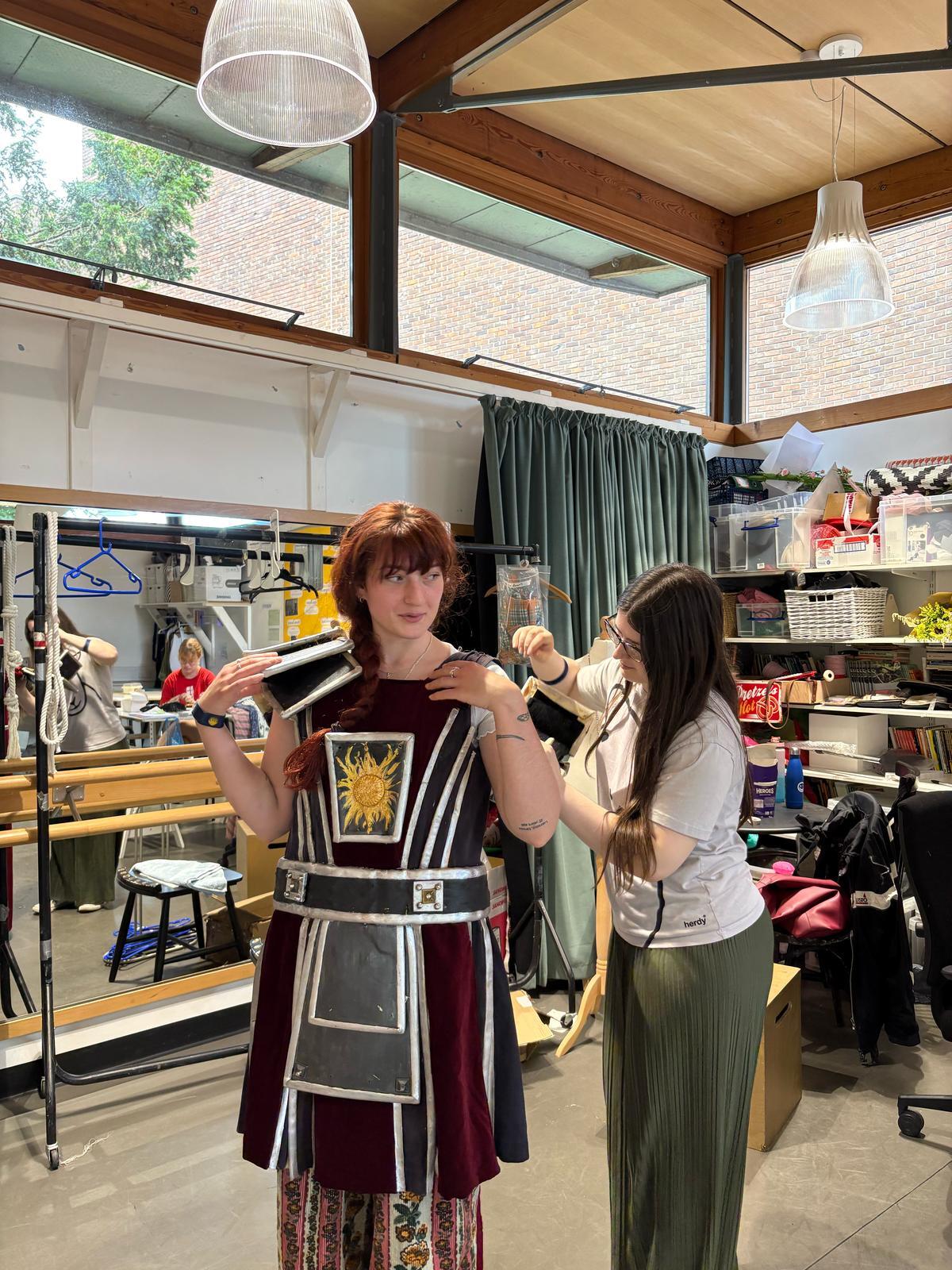 Woman with auburn hair holds burgundy and black geometric costume whilst another woman adjusts it in art studio with exposed beams.