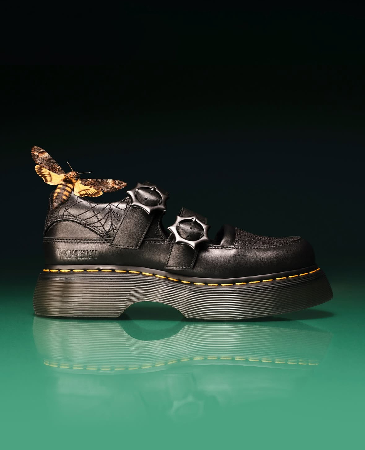Black leather platform shoe with metal buckles and patterned fabric accent, chunky ridged sole, against gradient green background.