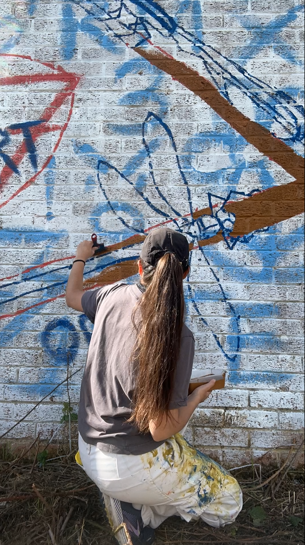 Person with long brown hair painting graffiti on white brick wall with blue, red and brown spray paint.
