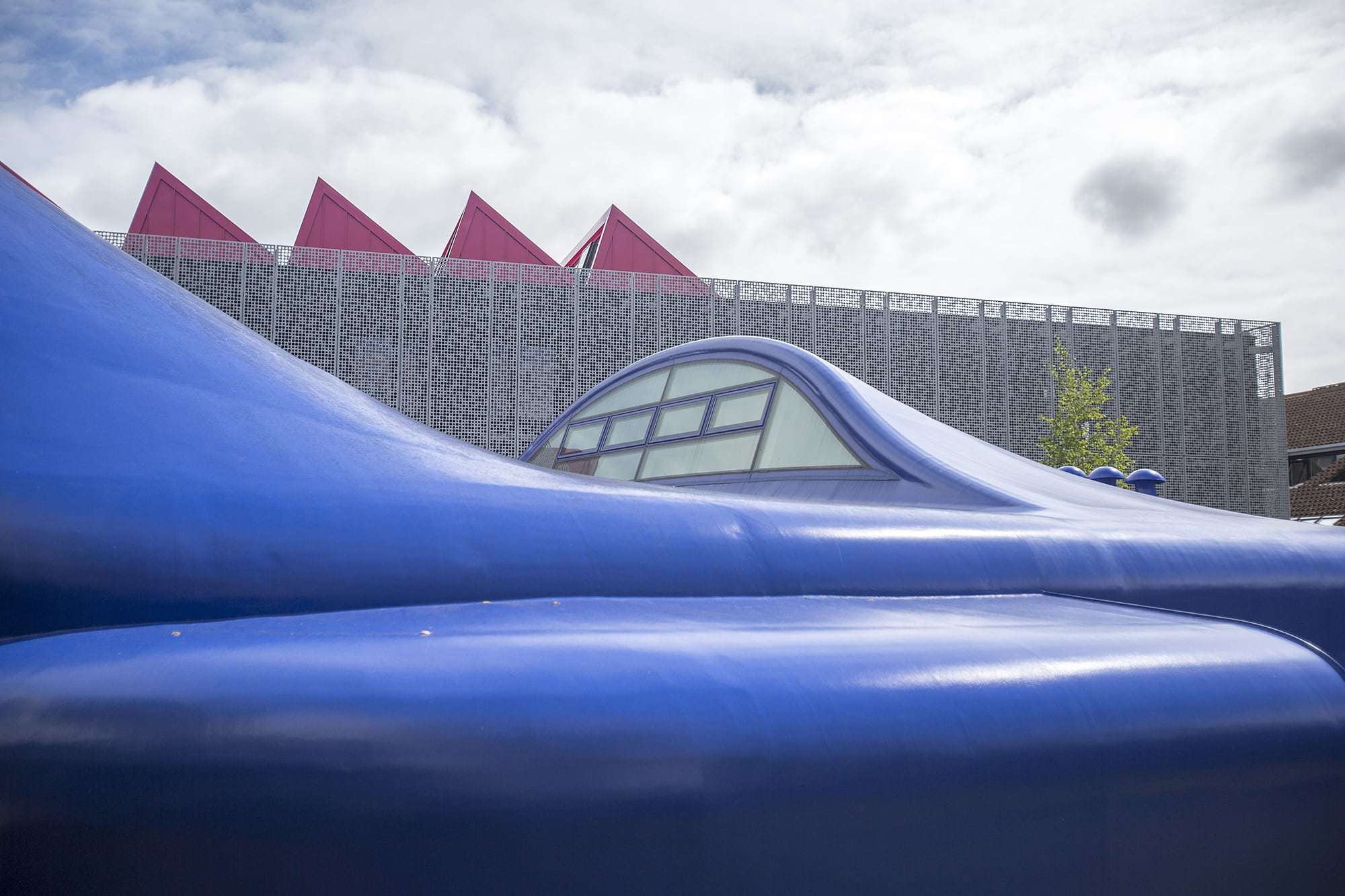A building with sloping blue rooftops in the foreground. Another large university building with metal mesh exterior and pink spiked rooftop stands in the background.