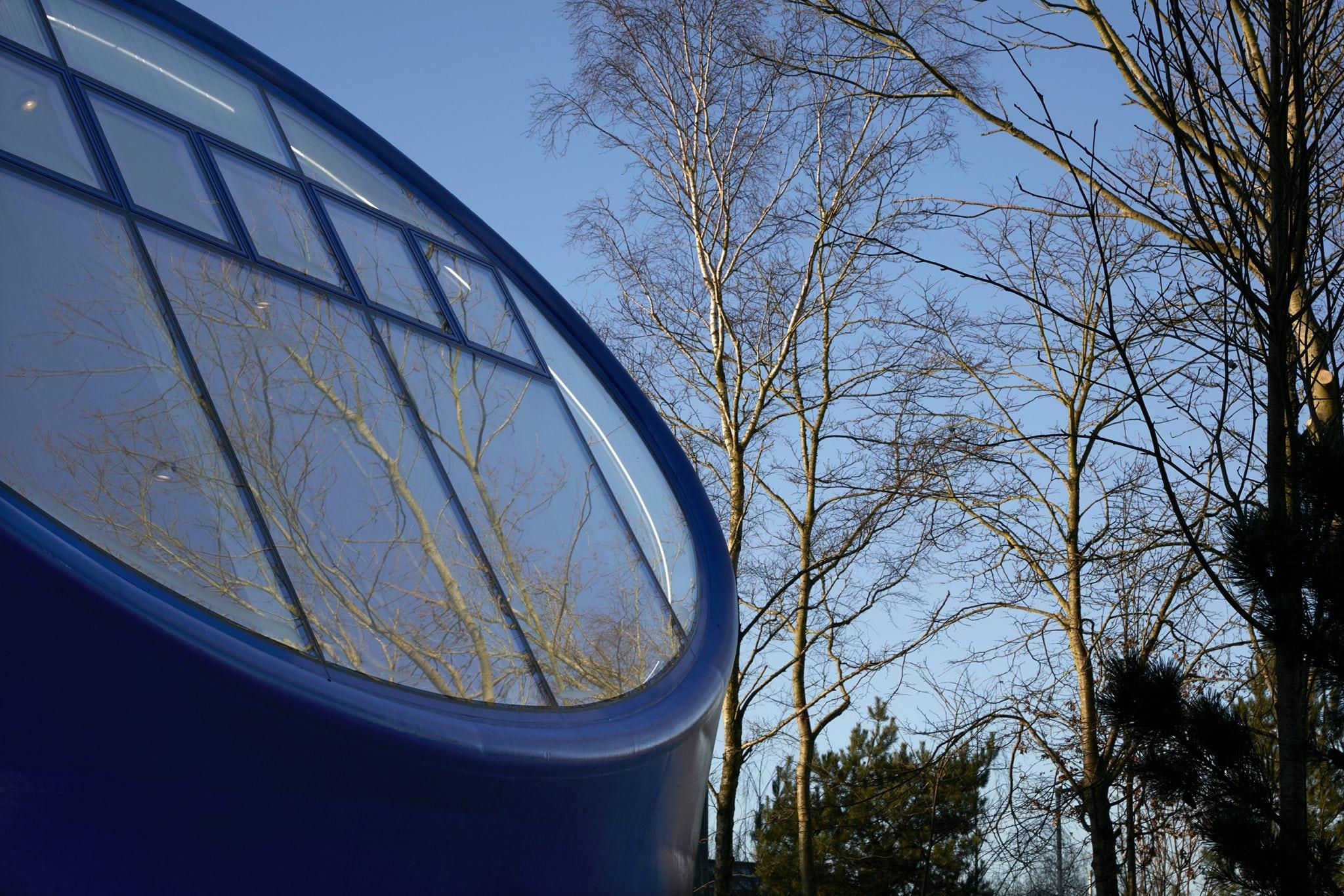Several windows fitting into a circular part of a building's roof, with trees standing on the right, reflected in the windows.