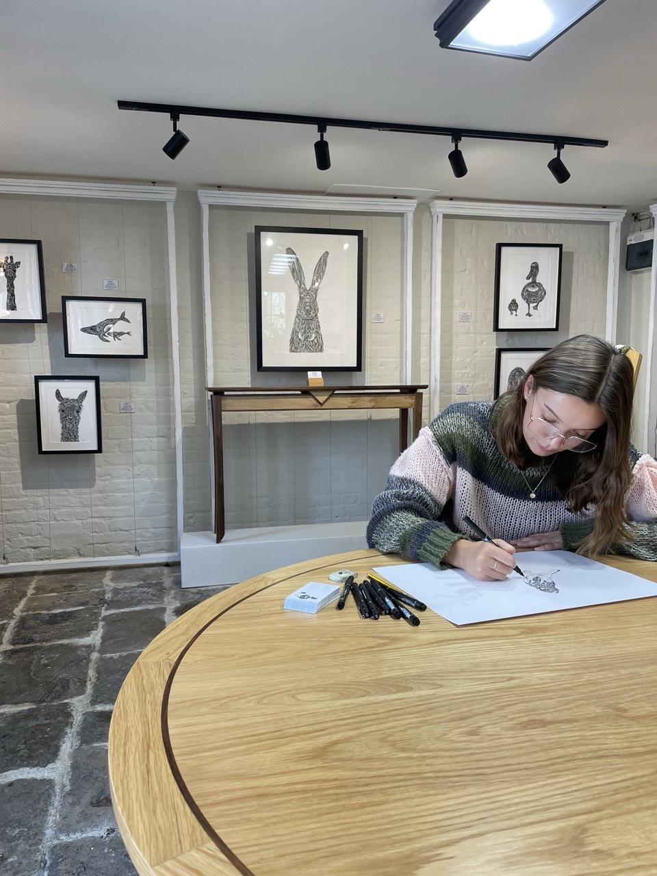 Pia Elliot drawing on a wooden table at her Intricate Wilderness gallery, wearing a cosy sweater and glasses, artwork framed in the background.