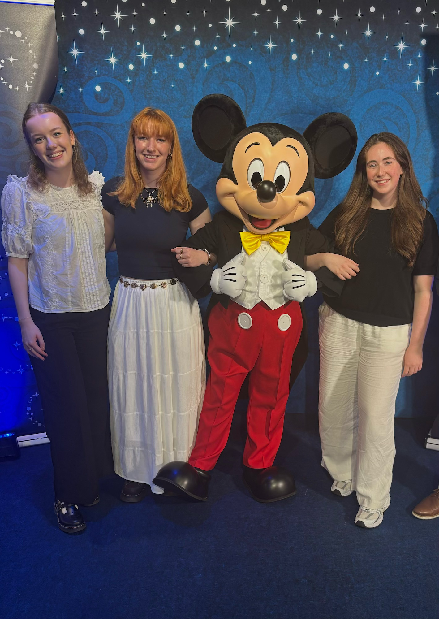 Three young people posing with Mickey Mouse character against starry blue backdrop with twinkling lights.