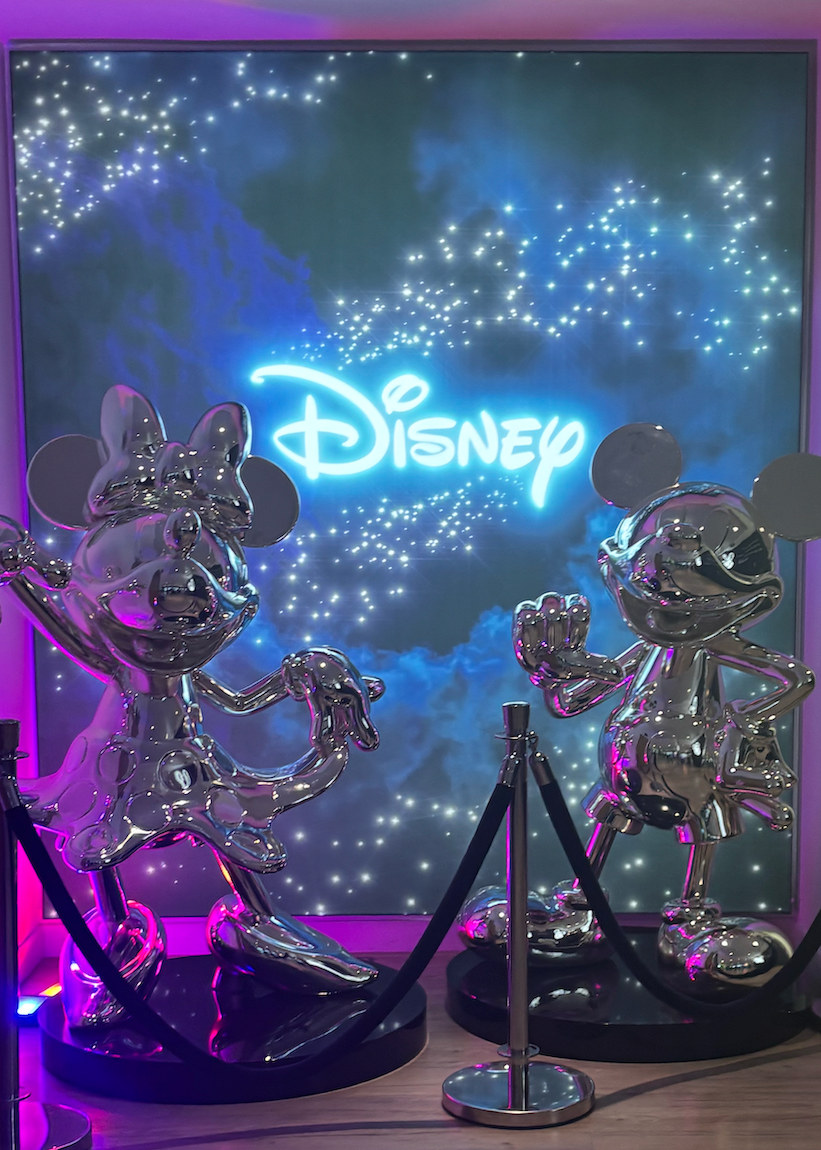 Silver Mickey and Minnie Mouse statues behind rope barrier, blue starry Disney logo backdrop with purple lighting effects.