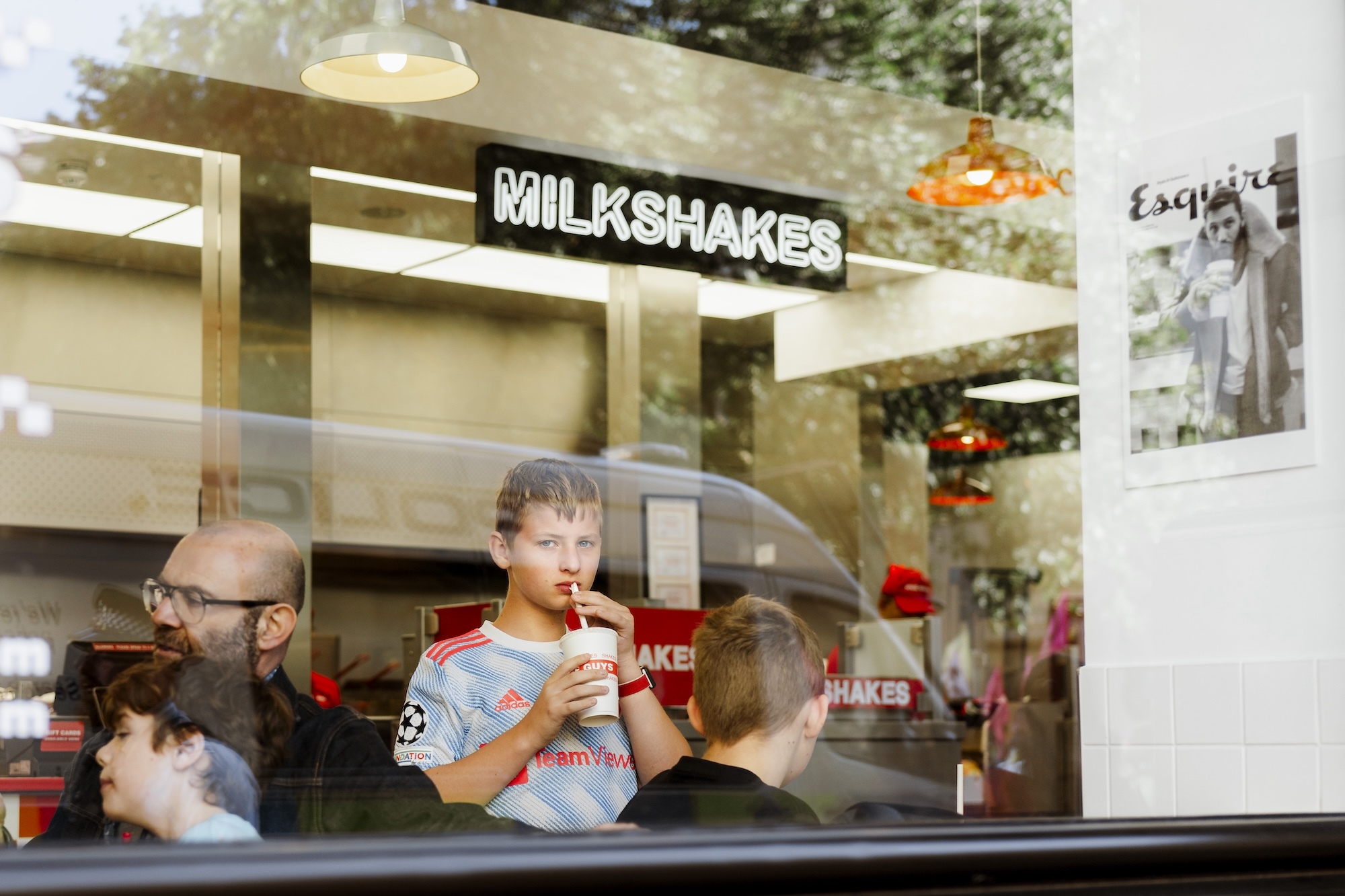 A young person looking at the camera through the window of a Five Guys restaurant sipping at a drink. A police van is visible in the reflection in the window.