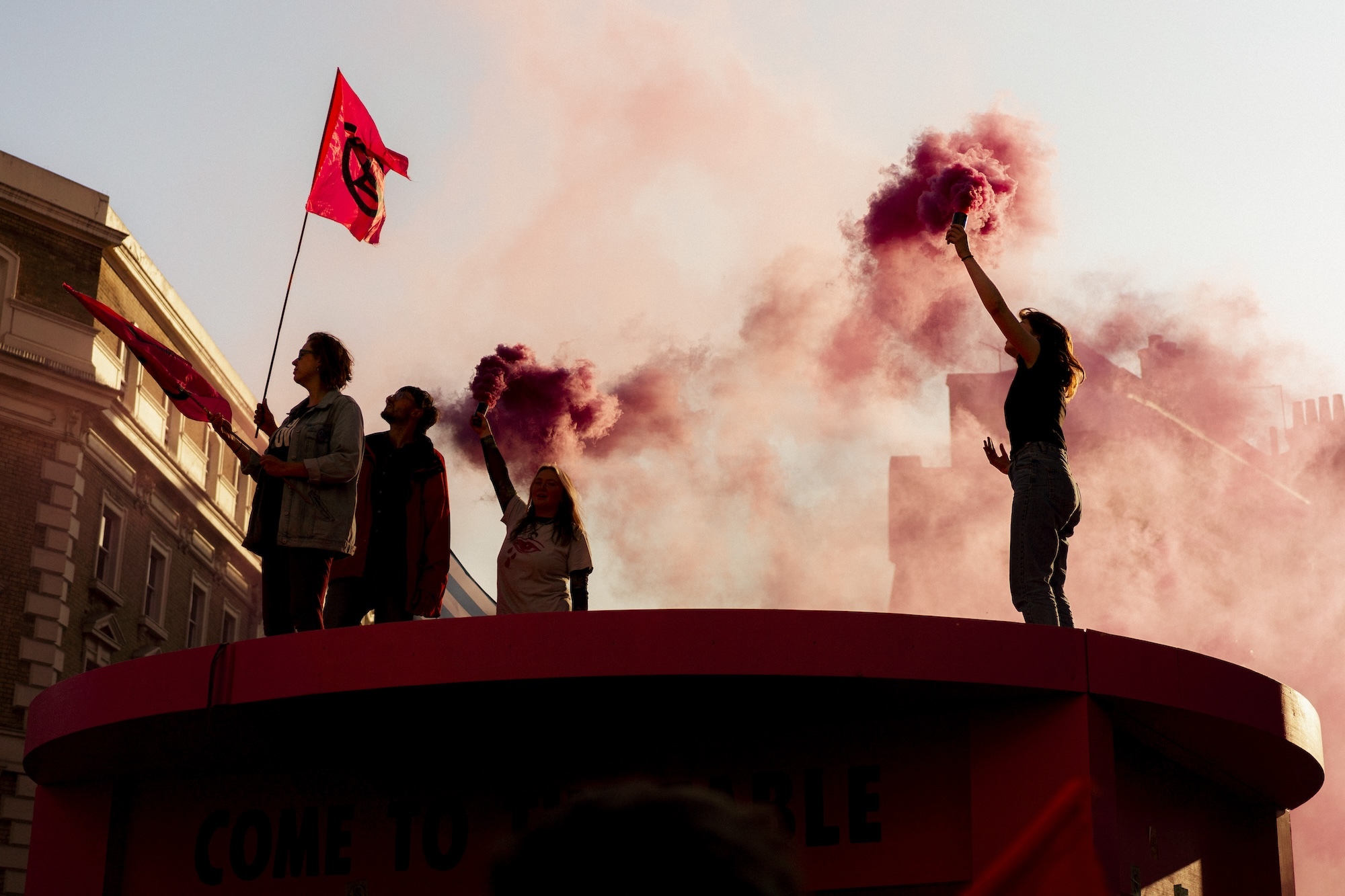 Four people on top of a roof waving red flags and raising canisters with red smoke billowing out.