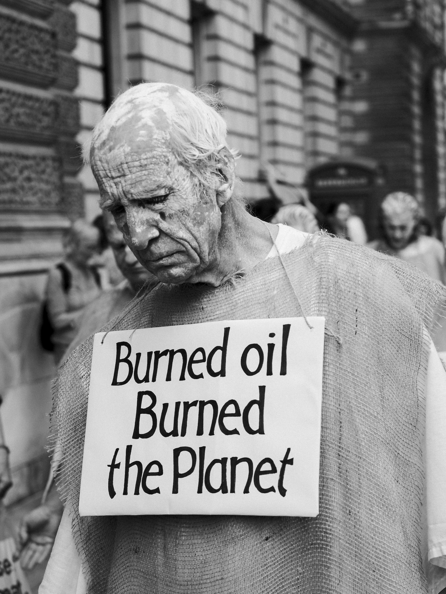 An older person with paint on their face walks through a street looking towards the ground, wearing a sign around their neck with text reading "Burned oil Burned the Planet".