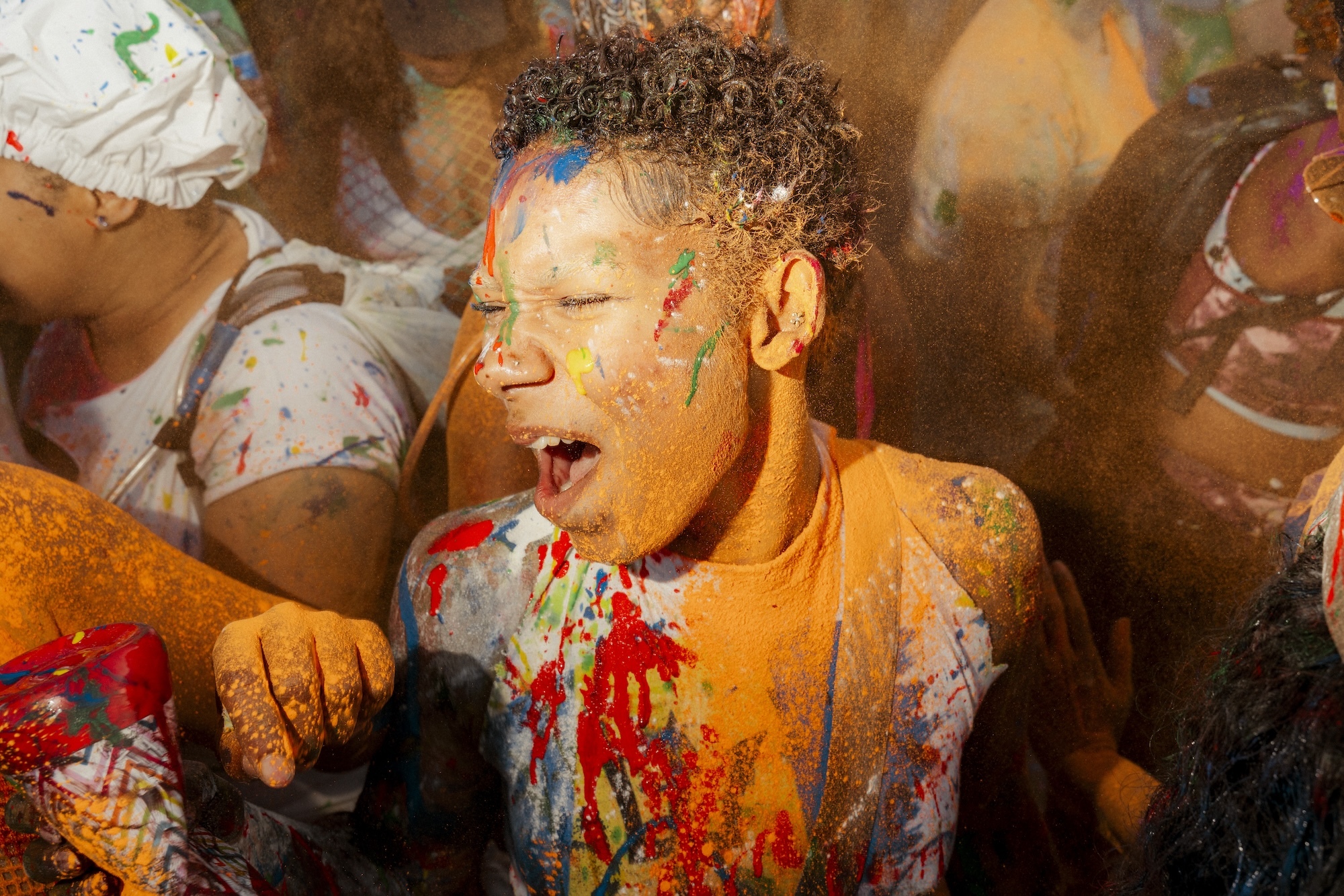 Close-up of a person in a crowd with their eyes screwed shut but their mouth open covered in orange paint.