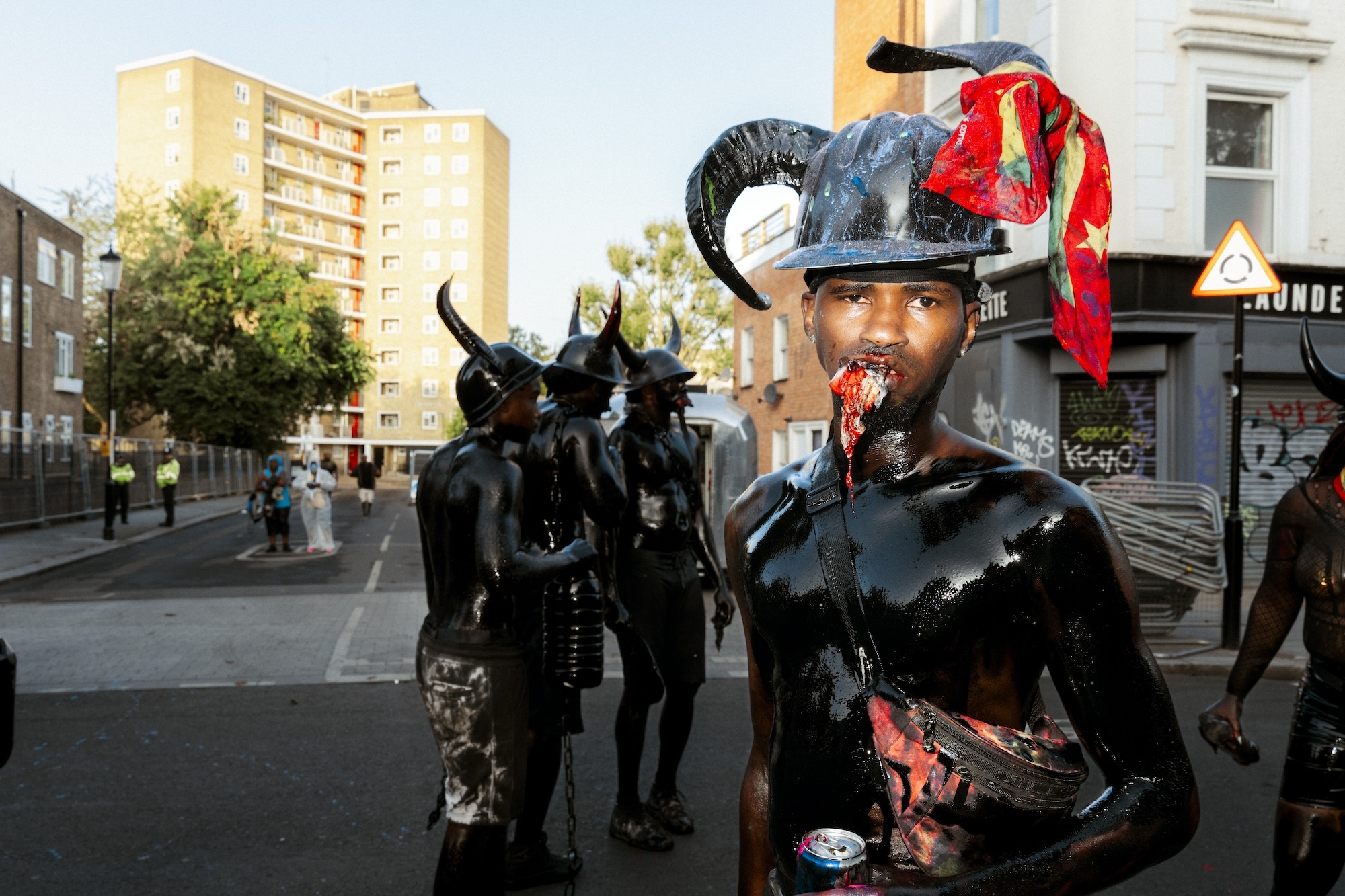 A shirtless man covered in oil chewing an object while wearing a black helmet with horns on it.