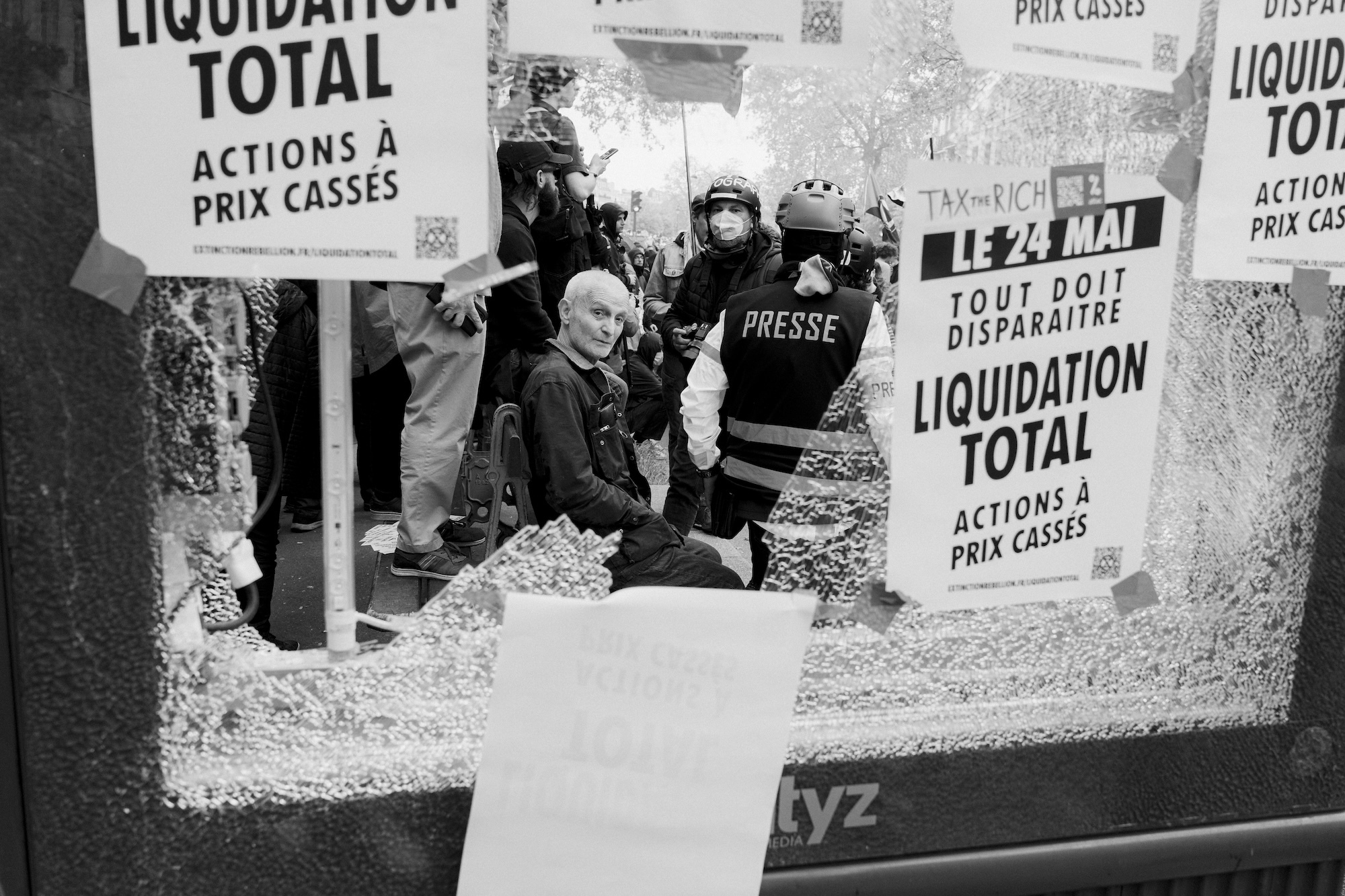 An older man sitting in the street surrounded by police, viewed through the glass of a broken window, with various posts in French taped to the window.