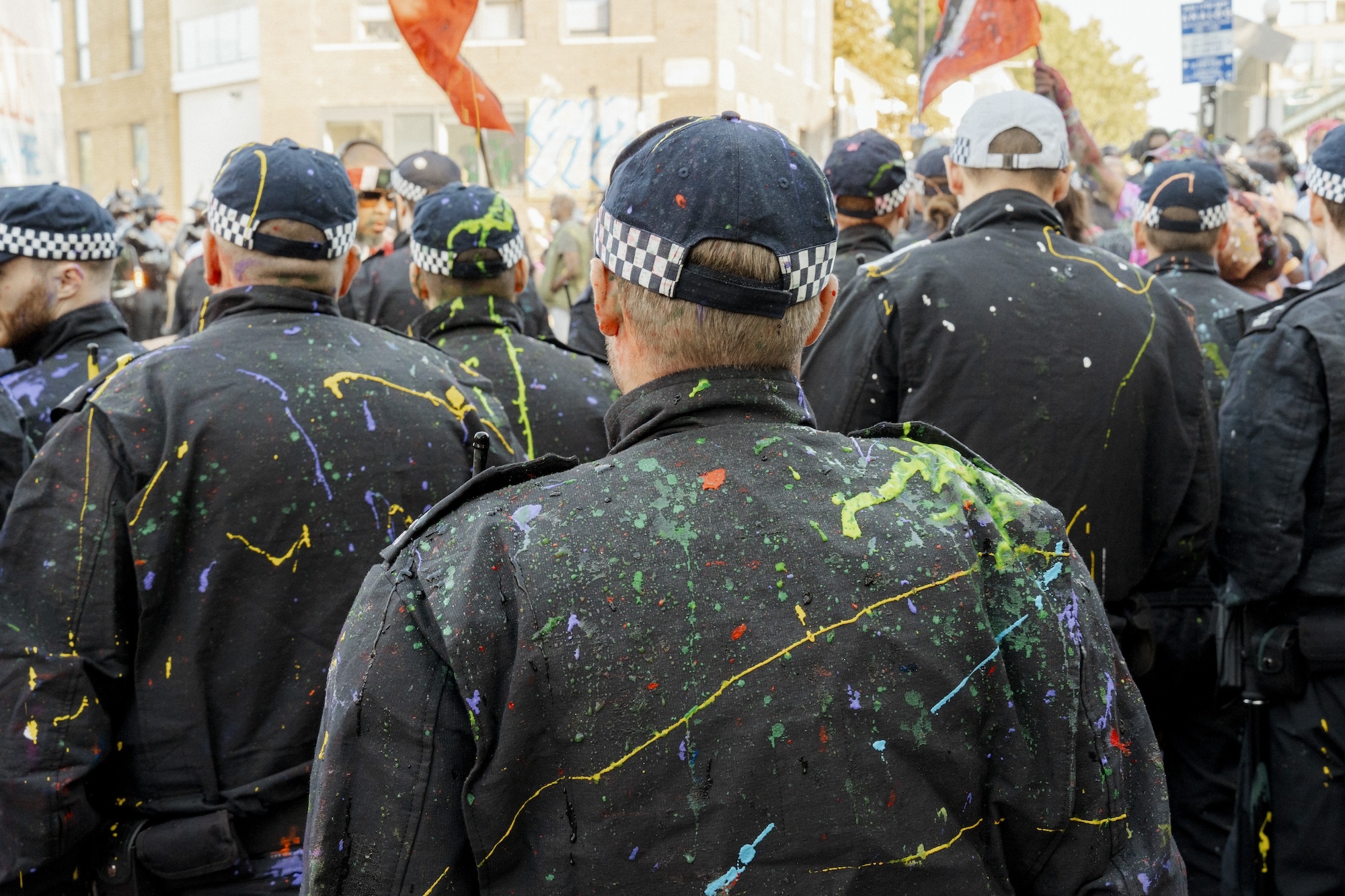 A crowd of policemen viewed from the back wearing black jackets covered in different coloured paints.