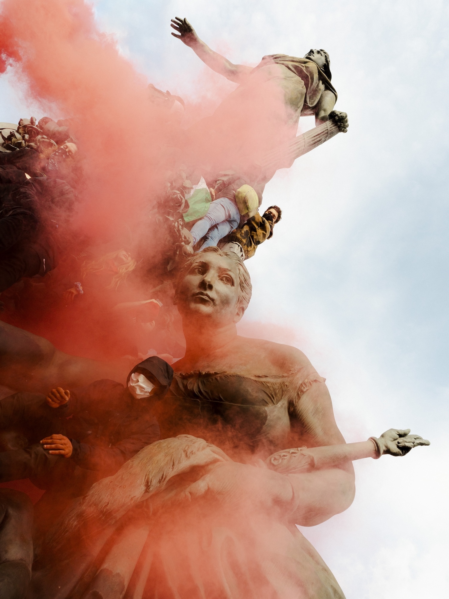 People wearing face masks taking shelter on statues surrounded by red-orange smoke during protests in Paris.