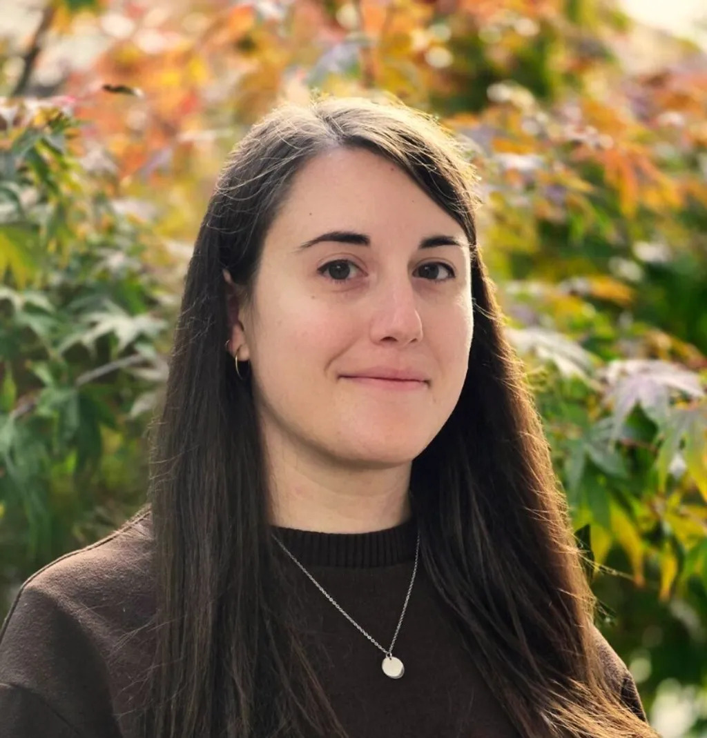 Woman with long dark hair wearing black top and pendant necklace, smiling at camera with autumn foliage background.