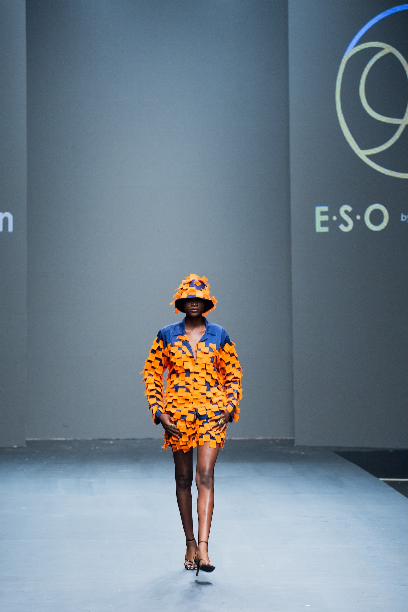 Model walks runway wearing orange patterned jacket and matching bucket hat over blue top, grey backdrop with partial "ESO" text visible.