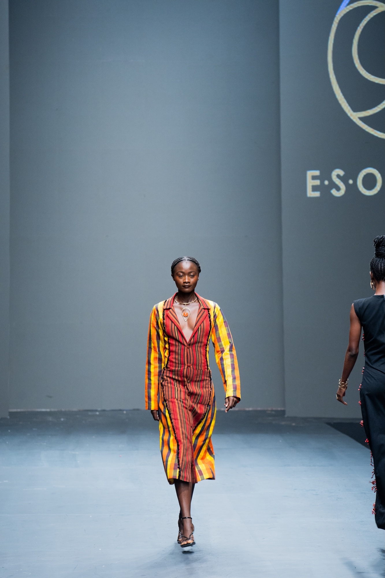 Black woman walking down fashion runway wearing colourful striped dress and matching coat. Grey backdrop with partial "E.S.O" text visible.