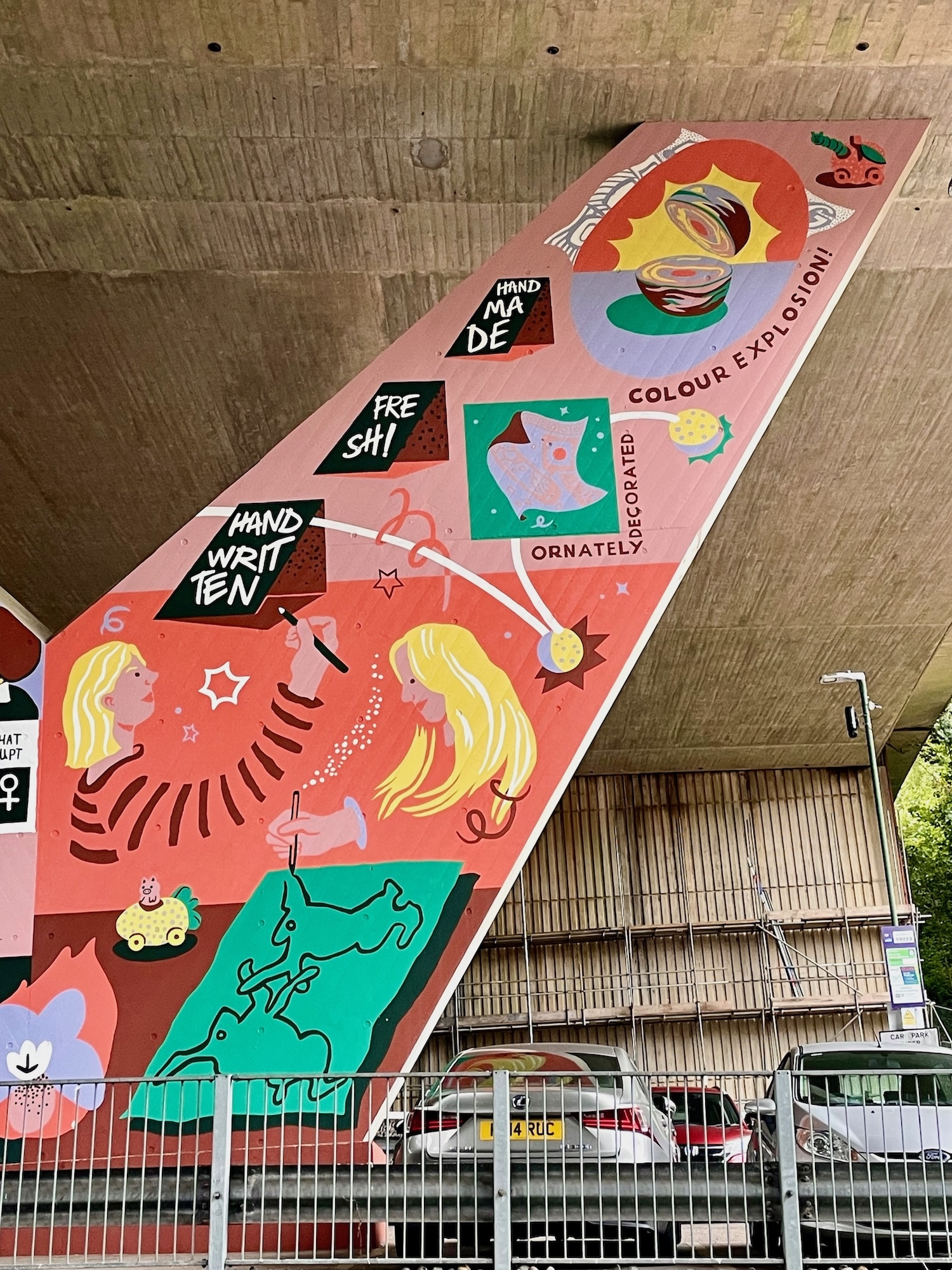 Part of a mural on a flyover pillar wall. Primarily red and pink showing illustrations of two people drawing, as well as a bath bomb split in half and various items of text.