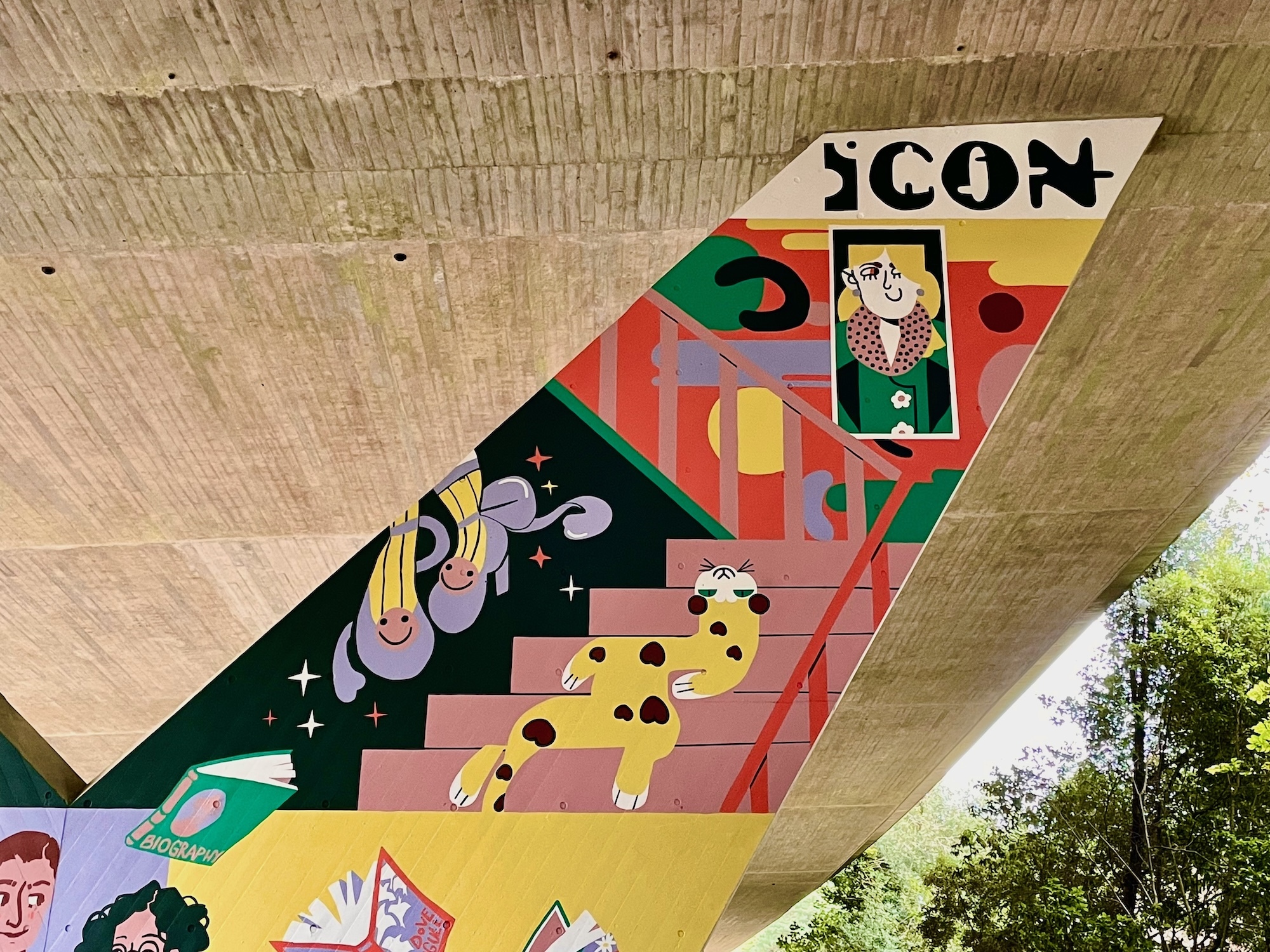 Part of a mural on a flyover pillar wall. A cat with heart-shaped spots lies on a stair case, which leads up to a multicoloured background and a woman's portrait in a frame on it. The word "Icon" appears at the top.
