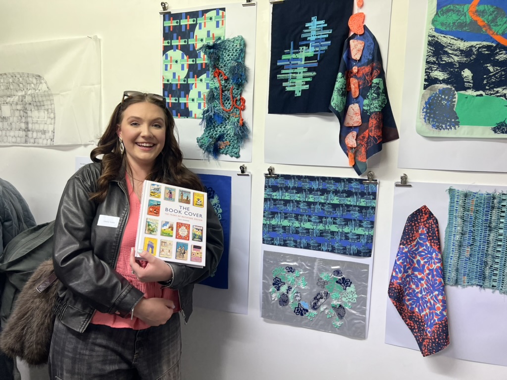 BA (Hons) Textiles Design student E Webber posing next to a display of textiles work on a white gallery wall.