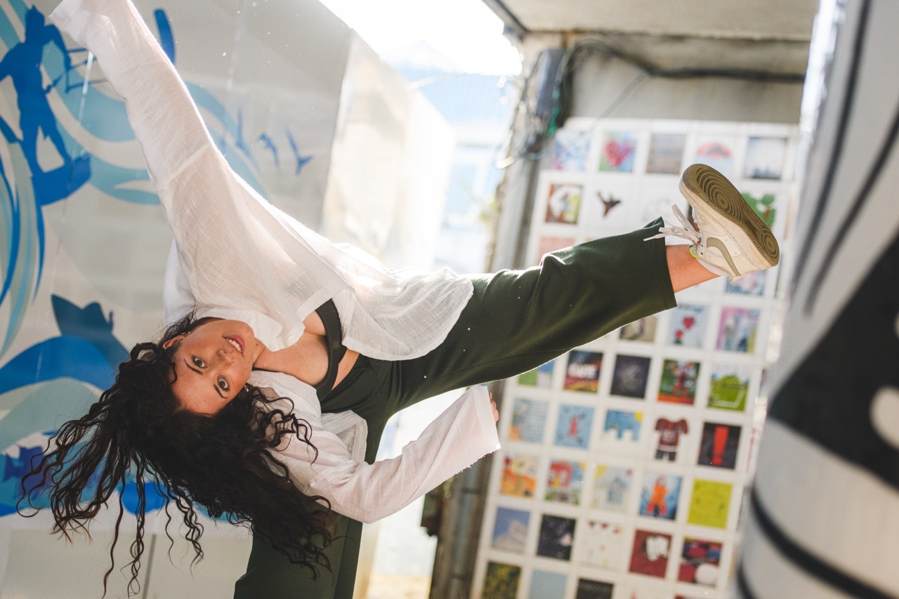 Woman with dark hair lying on floor in white shirt and green trousers, surrounded by colourful artwork on walls in studio space.