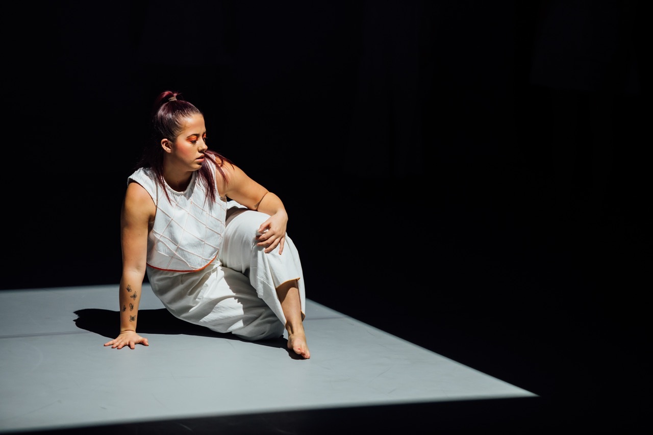 Woman in grey clothing sits on illuminated grey floor against black background, hand raised to face in contemplative pose.