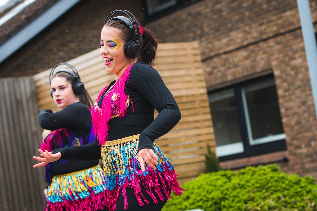 Two women dancing outdoors wearing black tops, colourful sequined skirts and headphones against brick building backdrop.