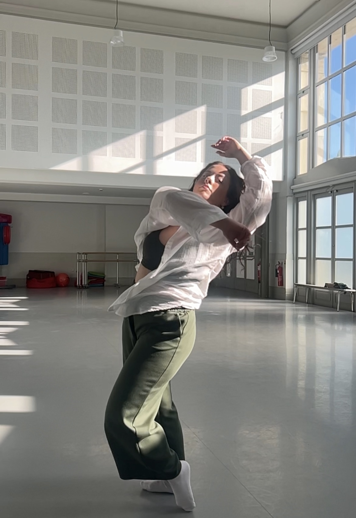 Digital illustration of figure in white shirt and green trousers dancing in sunlit studio with grey walls and large windows.