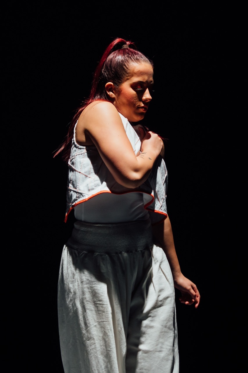 Woman in white dress with black corset-style bodice performing on stage against dark background, hair in ponytail with reddish tint.