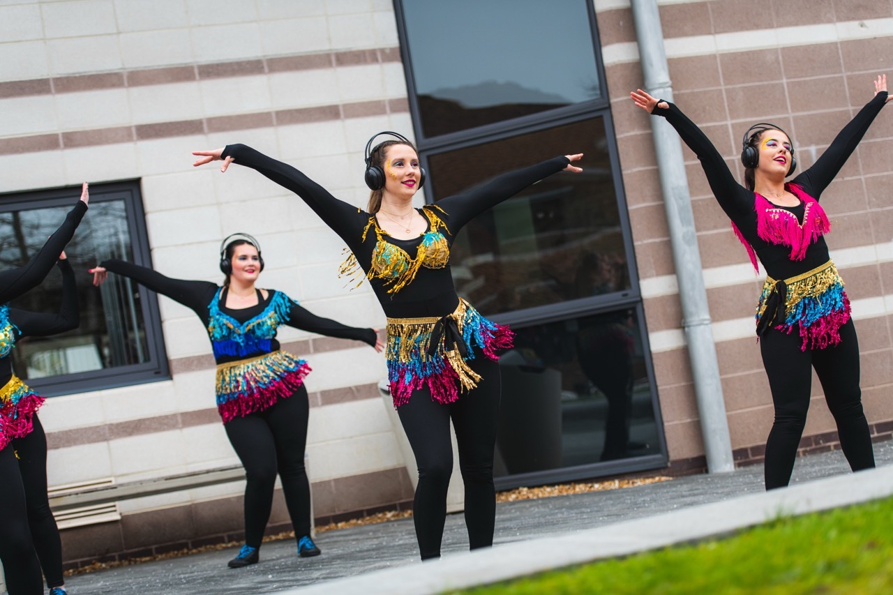 Three women in black leggings and colourful tops with arms raised, performing outside modern building with glass entrance.