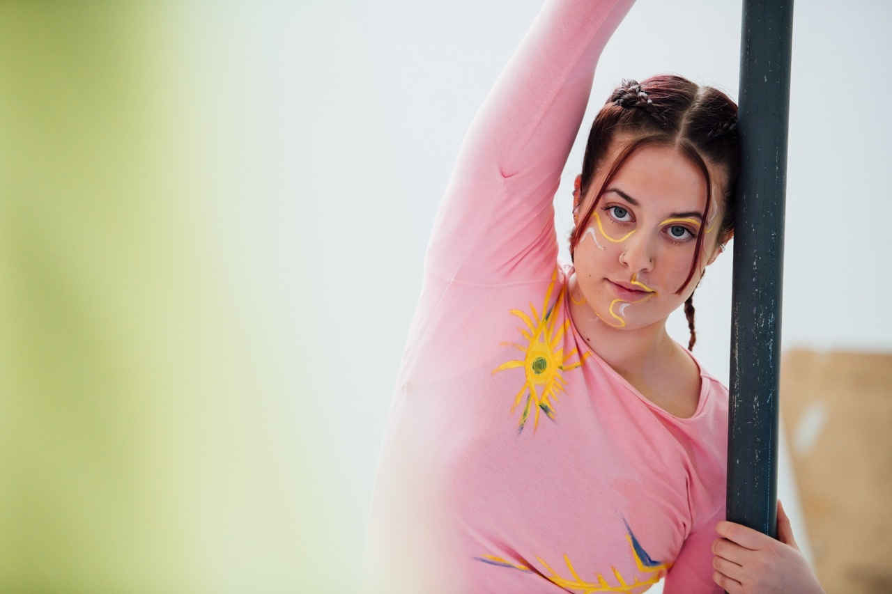 Woman in pink top with yellow flower in hair standing beside black vertical beam, colourful blurred background of green and pink.