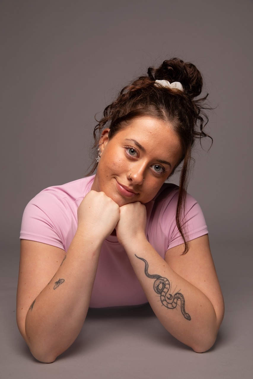 Woman with brown hair in messy bun wearing pink t-shirt, chin resting on hands, snake tattoo visible on forearm against grey background.
