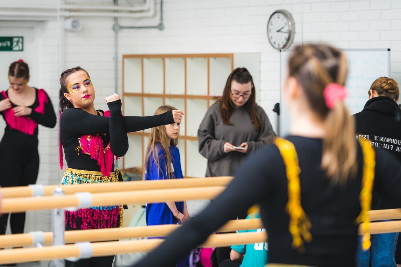 Dancers in black top and colourful skirt stretching at wooden barre, students in the background.