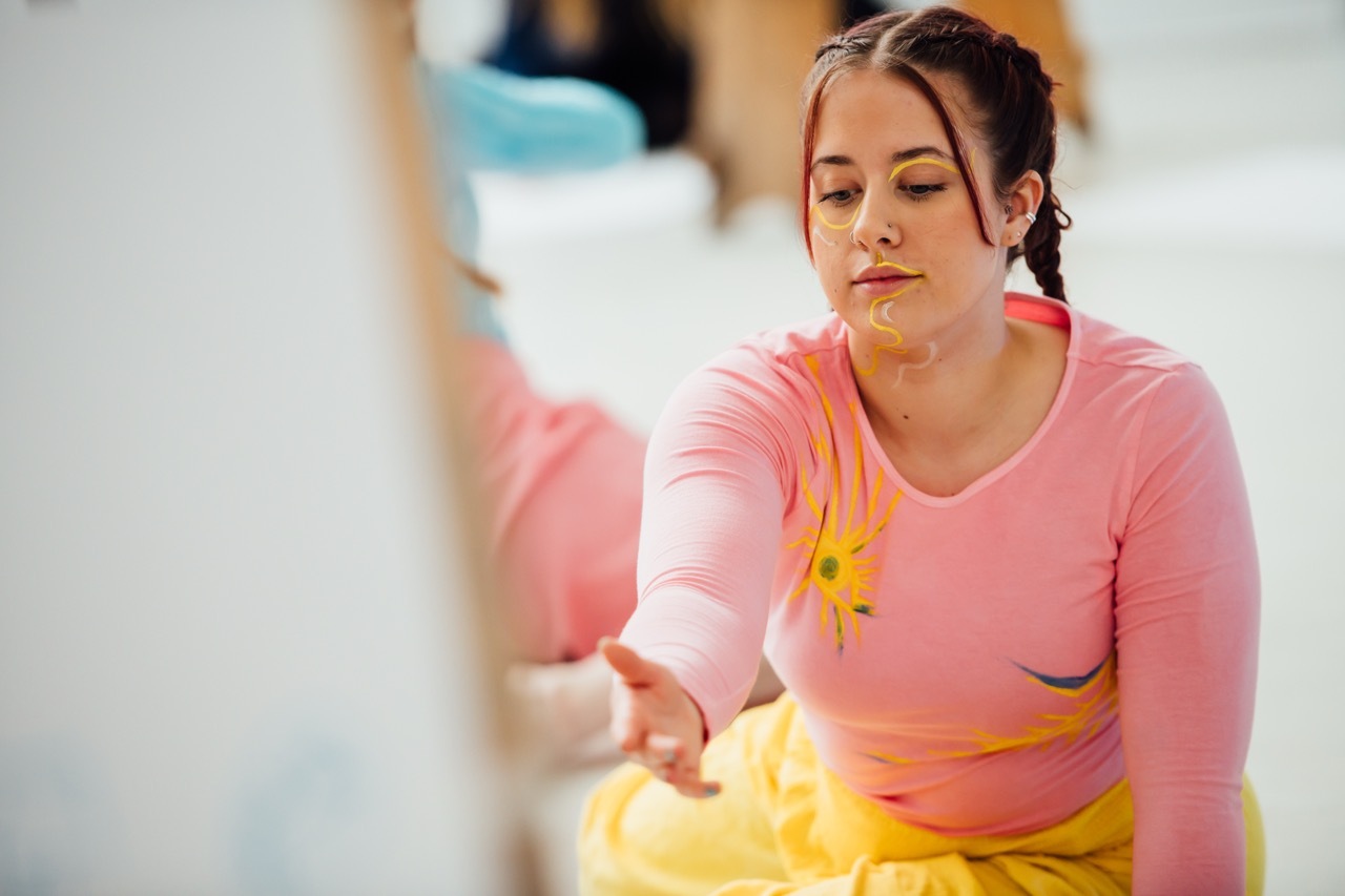 Woman in pink long-sleeved top and yellow skirt sitting, looking downward with hair in ponytail against blurred background.