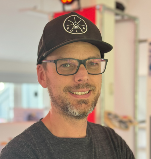Man wearing black baseball cap with circular logo, black-rimmed glasses, and grey beard smiling in bright indoor space.