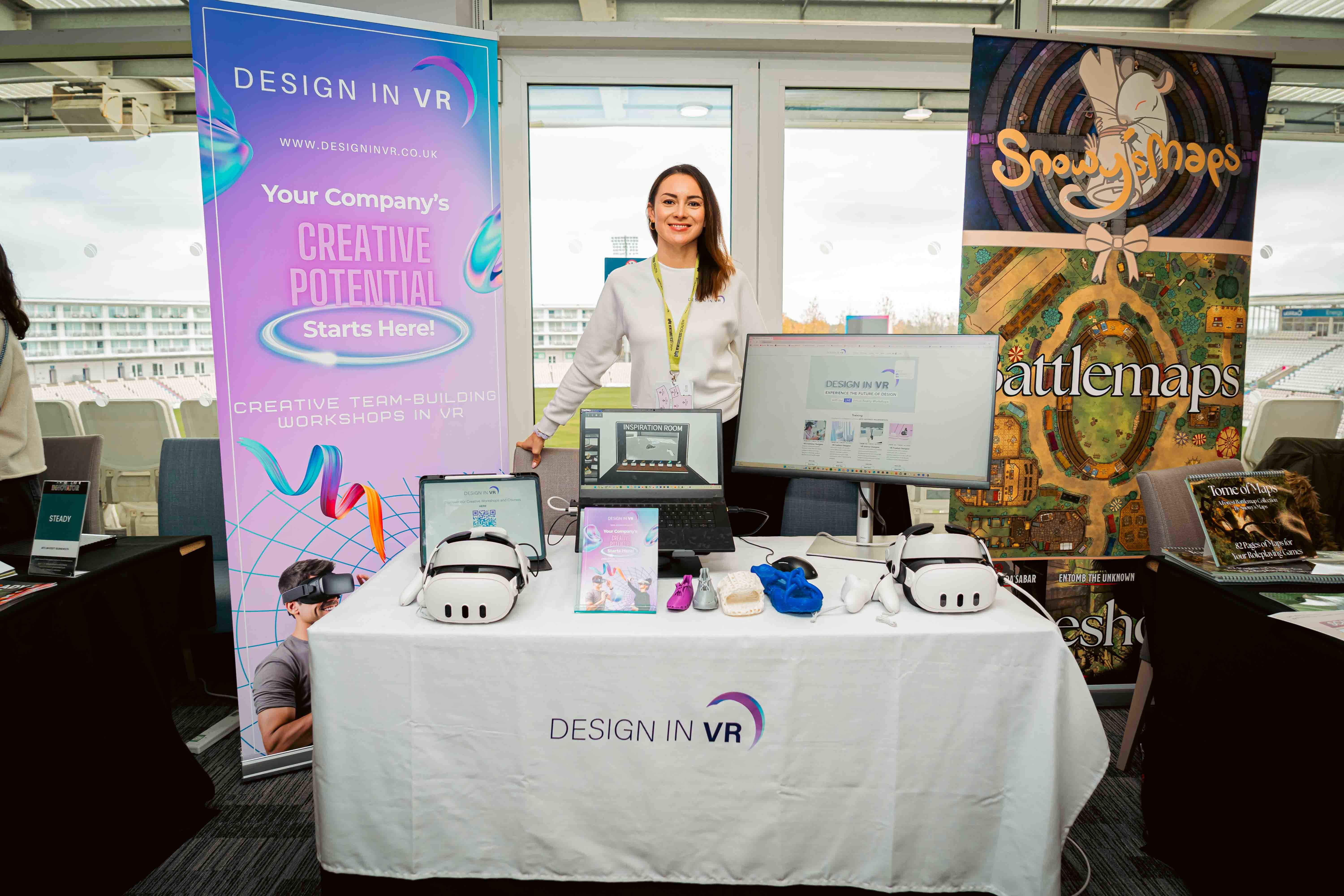 E Savosina at her stall for Design in VR at Venturefest South 2024. She is standing behind a table with computer monitors and VR equipment and a banner for Design in VR to the left-hand side.