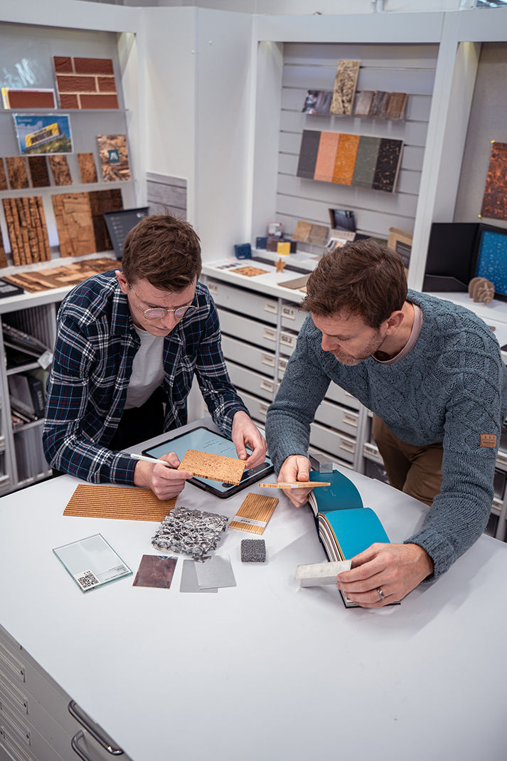 Two men examining material samples at white desk in showroom with wood, tile, and colour samples displayed on walls and shelves.