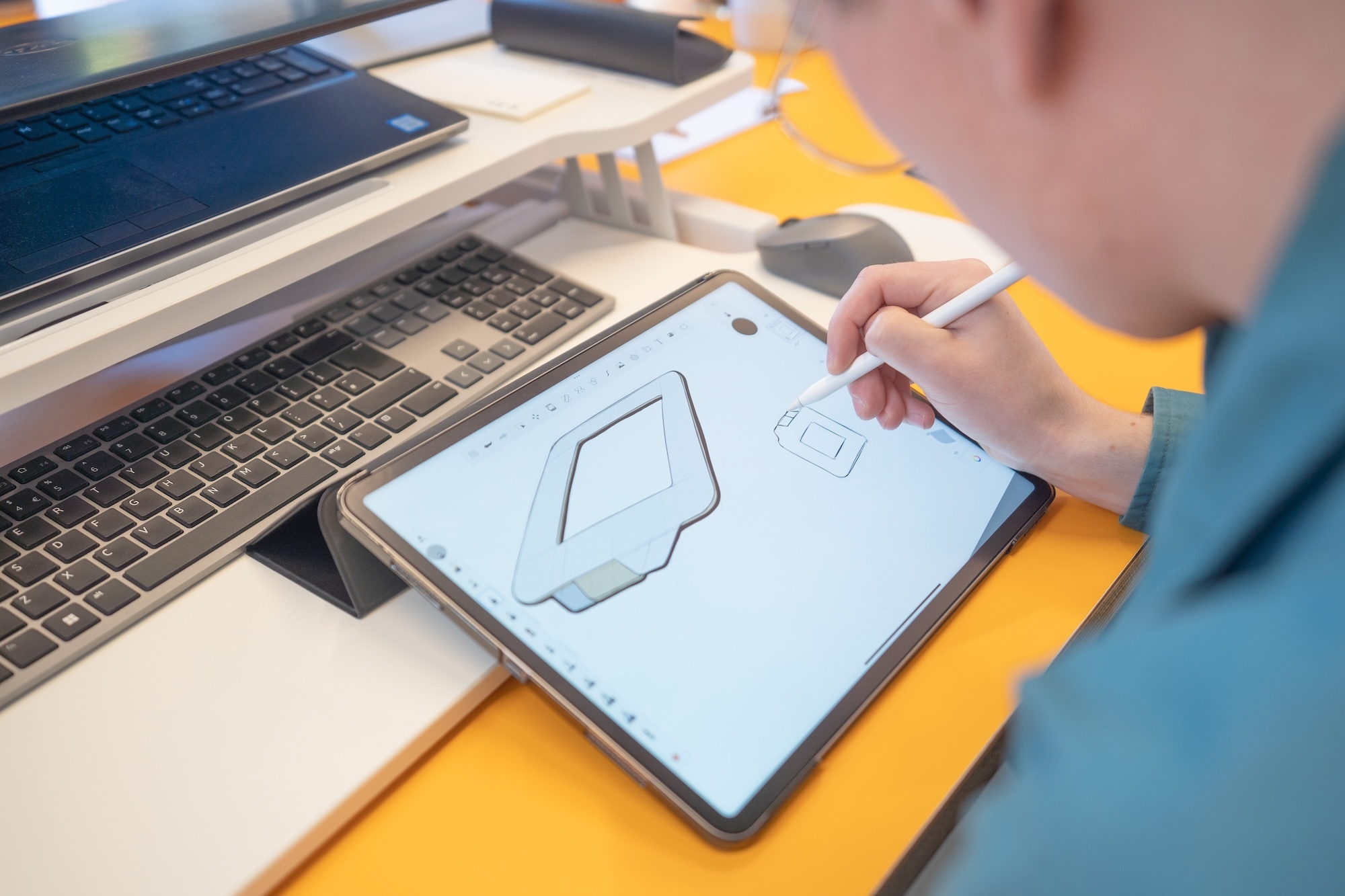 Close-up of a tablet with design software open on the screen. A person is sketching next to a design with a stylus.