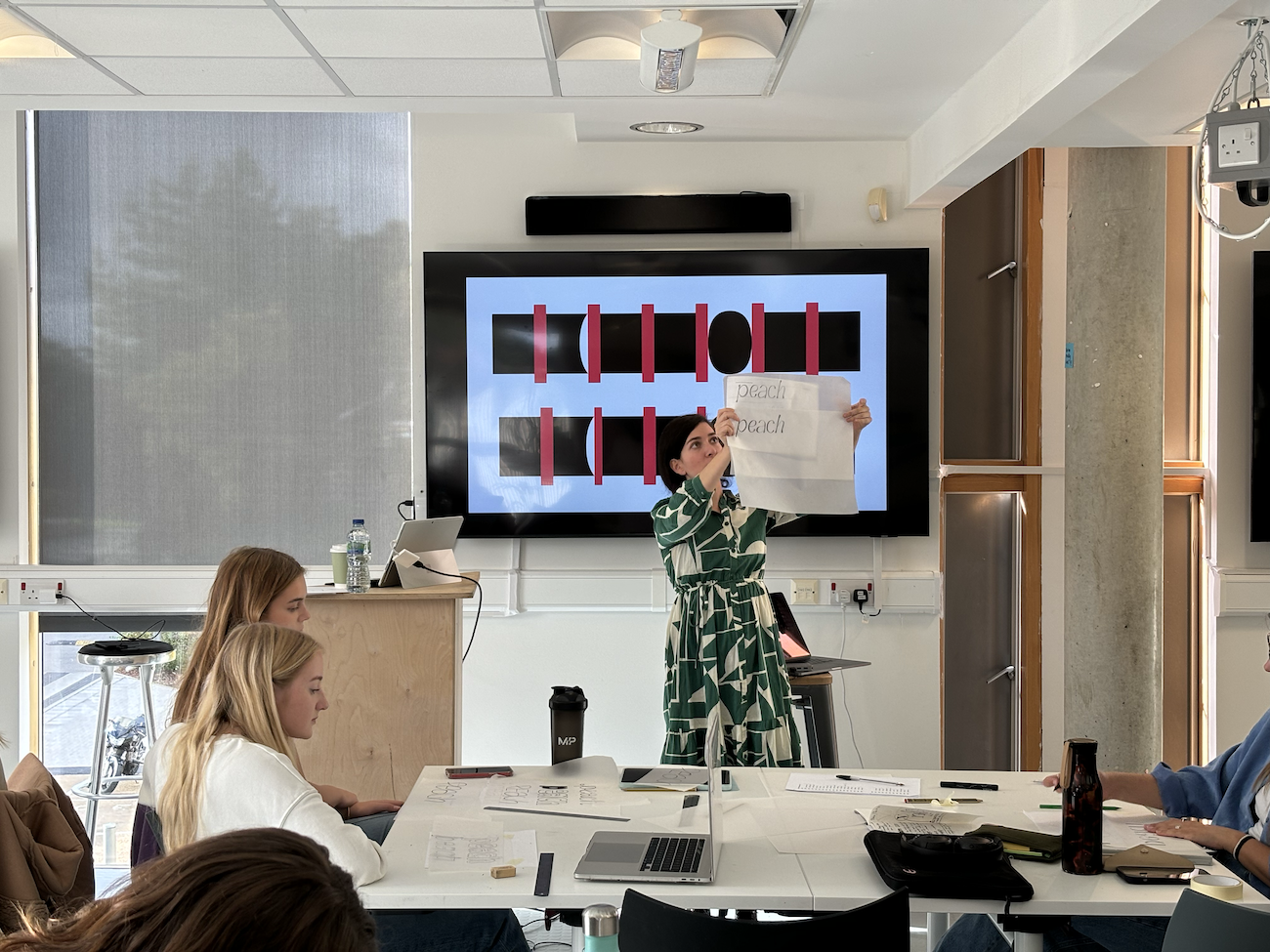 Eleni Beveratou stands in front of a table with BA (Hons) Graphic Design students sitting. Eleni holds up a piece of paper with the word 'peach' written twice in an italic-style font.