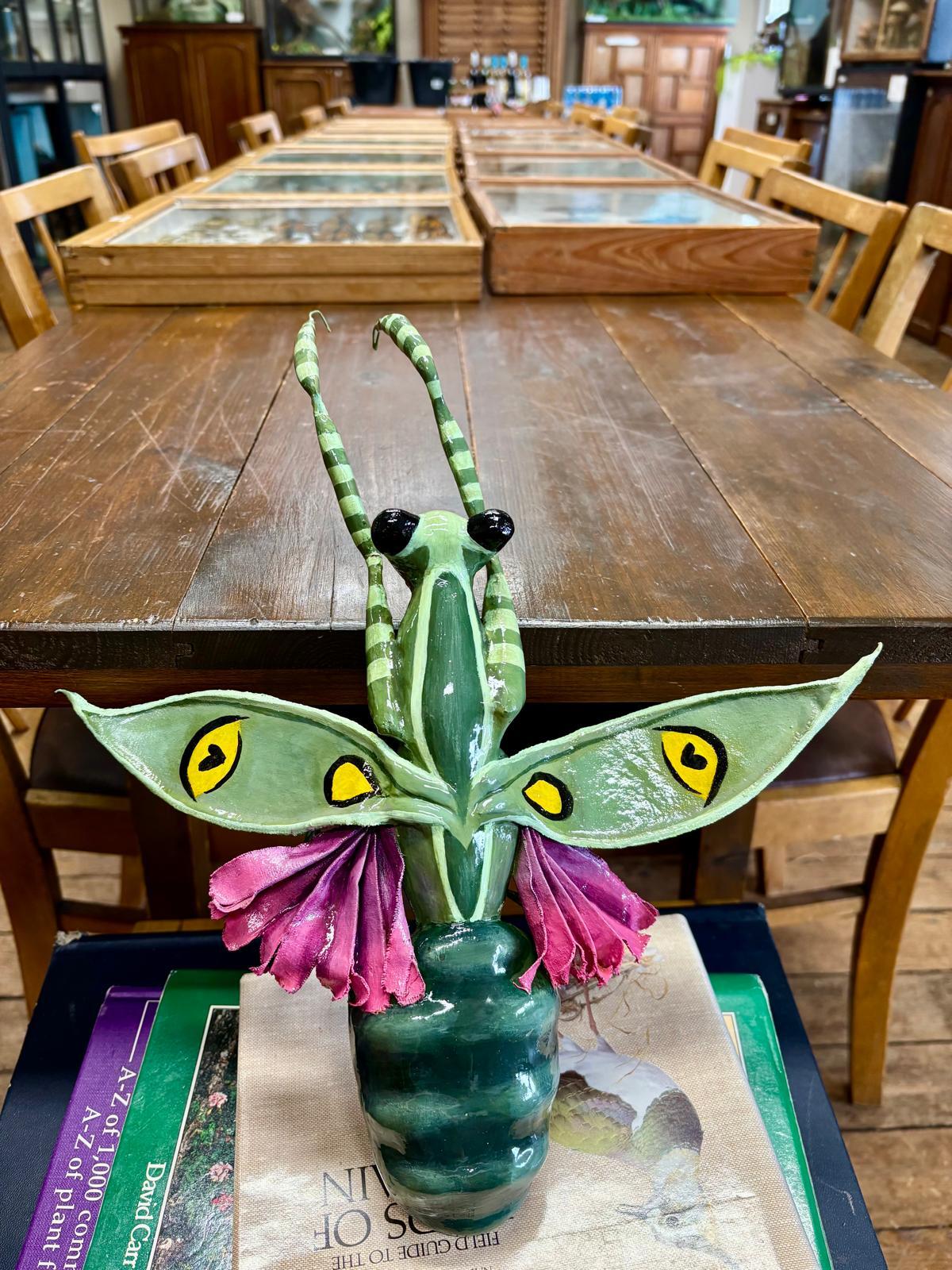 Green sculptural figure with yellow eyes, purple wing-like elements, and striped antennae on wooden table in rustic interior setting.